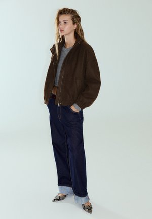 Jeune femme debout, les mains dans les poches, portant une veste marron, un t-shirt gris, un jean foncé retroussé et des chaussures pointues à motif peau de serpent.