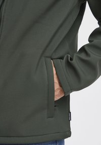 Dark green softshell jacket with a zip front, featuring side pockets and a textured finish. Includes branding tag on the lower edge.