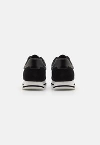 Chaussures de sport noires avec un dessus en daim et des accents brillants, présentant une semelle intermédiaire blanche et des détails noirs le long de la semelle et du talon.