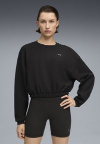 Sort cropped sweatshirt med elastisk nederkant, der har et diskret gråt logo. Kombineret med sorte tætsiddende shorts, begge i glat stof.