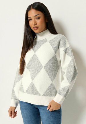 Vrouw met lang donker haar, die een wit-grijs argyle patroon trui en een blauwe spijkerbroek draagt, staand tegen een effen achtergrond.