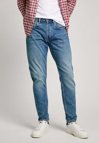 Blue denim jeans with a faded wash, straight cut, and five-pocket design, paired with white sneakers and a checkered red shirt.