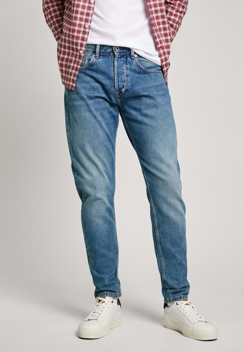 Jean en denim bleu avec un délavage, coupe droite, et design à cinq poches, associés à des baskets blanches et une chemise à carreaux rouges.
