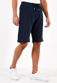 Pantaloni corti in cotone blu navy con vita elastica e coulisse, dotati di tasche laterali e vestibilità rilassata, abbinati a sneakers bianche.