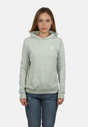 Frau mit langen braunen Haaren trägt einen hellgrauen Kapuzenpullover mit kleinem Herzlogo und blaue Jeans, steht vor einem weißen Hintergrund.