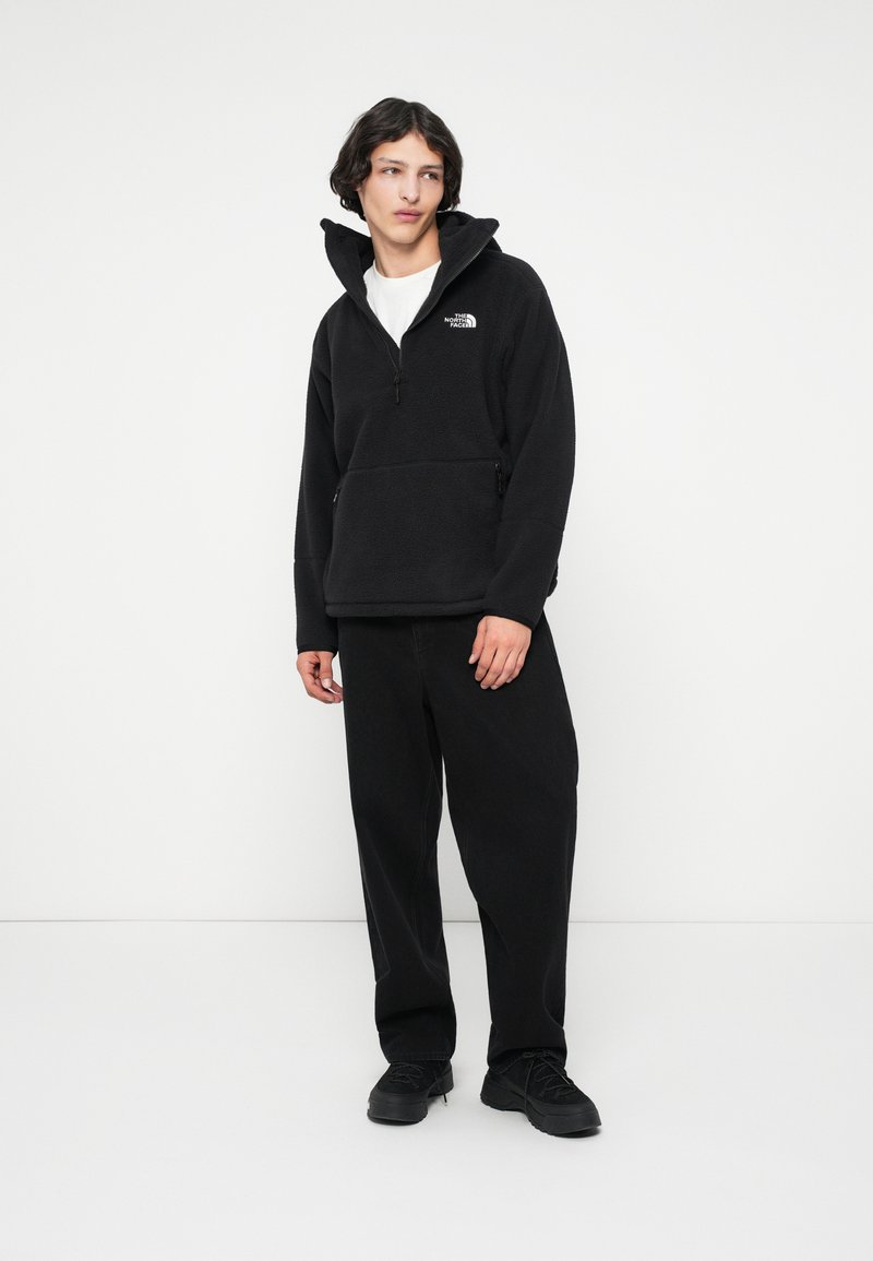 Junger Mann in einer schwarzen Fleecejacke mit dem The North Face-Logo, schwarzen Hosen und schwarzen Schuhen, steht vor einem schlichten weißen Hintergrund.