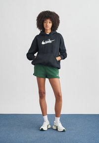 Μαύρο φούτερ Nike με λευκό λογότυπο, πράσινη βερμούδα, λευκές κάλτσες και ανοιχτόχρωμα αθλητικά παπούτσια. Απαλή υφή, χαλαρή εφαρμογή, στυλ casual αθλητικής ένδυσης.