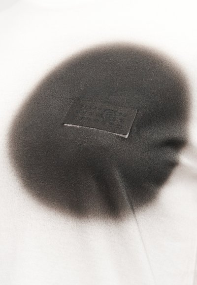 Μαύρο κυκλικό μοτίβο με ψεκασμό, με μια μικρή ραμμένη ορθογώνια ετικέτα υφάσματος σε φωτεινό φόντο υφάσματος.