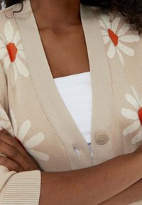 Kobieta w beżowym kardiganie z białymi i pomarańczowymi wzorami stokrotek, z założonymi na piersi rękami, w białej bluzce.