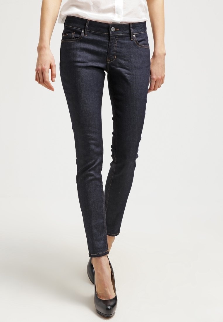 Jeans ajustados de mezclilla en azul oscuro, con bolsillos frontales, costuras sutiles y un corte Slim. Combinados con zapatos de tacón negros y una camisa blanca.