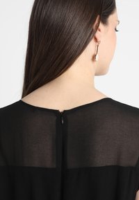 Schwarzes Kleid mit einer durchsichtigen oberen Schicht und einem festen unteren Abschnitt, mit einem Reißverschluss auf der Rückseite und strukturierten Stoffdetails.