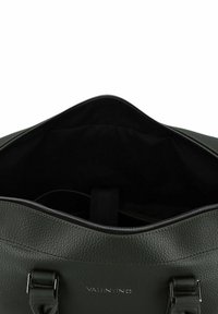 Sac à main en cuir vert, présentant une finition texturée, une fermeture zippée et une poche intérieure, avec le logo de la marque visible sur le devant.