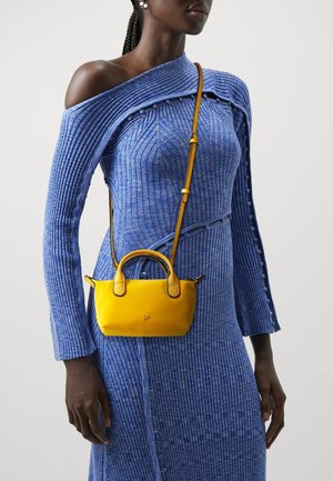 Vrouw die een blauw geribd off-shoulder jurkje draagt en een kleine gele crossbody-handtas met bovenhandvatten bij zich heeft.