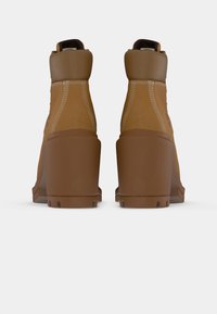 Un paio di stivali ankle in pelle marrone con tacchi spessi e cuciture bianche visibili, visti da dietro su uno sfondo bianco.