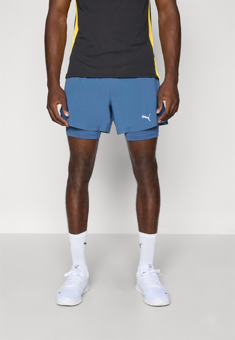 Pantalones cortos deportivos azules hechos de material ligero, con un logo blanco de Puma, combinados con calcetines blancos y zapatillas.