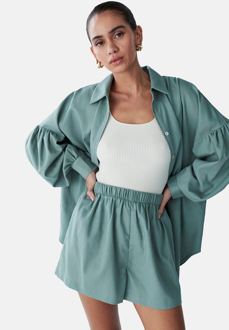 Světlě zelená oversized košile s nabíranými rukávy, spárovaná s bílým ribbovaným topem a odpovídajícími světlě zelenými kraťasy s elastickým pasem.