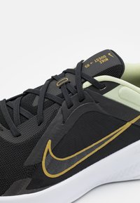 Baskets Nike noires avec une tige en mesh, des accents dorés et un col vert clair. Elles disposent de lacets, d'une languette rembourrée et d'une semelle blanche contrastée.