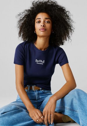 Kvinde med krøllet hår iført en marineblå "Red Bull Racing" t-shirt, lyseblå jeans og et brunt bælte, siddende med korslagte ben mod en grå baggrund.