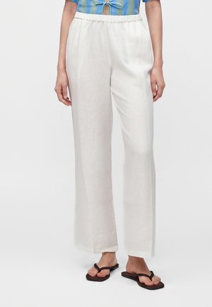 Vrouw draagt losse witte broek, een blauw overhemd met knoop vooraan en bruine teenslippers, staand tegen een effen achtergrond.
