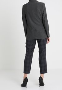 Blazer grigio su misura con una texture liscia e una sola apertura. Abbinato a pantaloni blu a scacchi e scarpe con tacco nere.