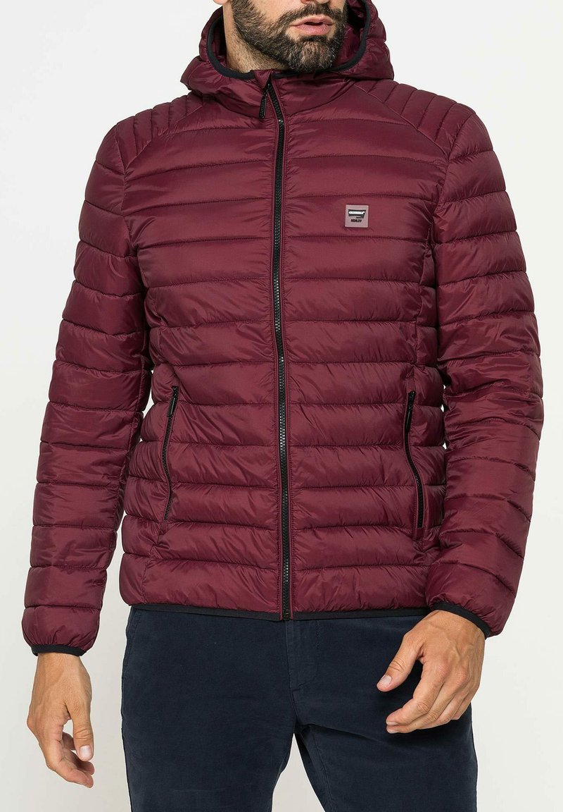 Giacca imbottita rossa con zip frontale, cappuccio e due tasche laterali. Presenta una texture trapuntata e una piccola patch con logo sul petto.