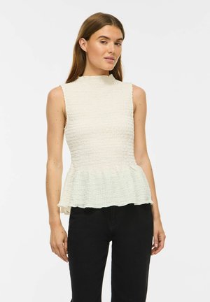 Woman wearing a sleeveless textured white peplum top and black pants standing against a plain white background.