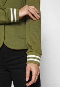 Giacca verde oliva con polsini a costine caratterizzati da strisce bianche. Design slim-fit con una texture liscia e tasche laterali.
