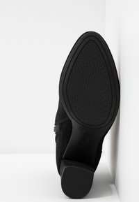 Chaussure en daim noir avec une semelle en caoutchouc texturée, dotée d'un talon incurvé et d'un détail élastique latéral. La semelle présente un motif embossé « SOSOS ».
