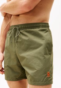 Șorturi de baie de culoare verde oliv, fabricate dintr-un material ușor. Au o talie elastică cu șnururi de strângere și un logo portocaliu pe piciorul inferior.