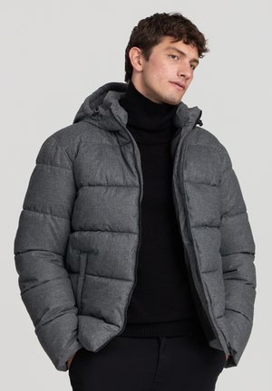 Giacca a vento grigia con cappuccio, caratterizzata da un design trapuntato, zip frontale e due tasche laterali. Indossata sopra un dolcevita nero.