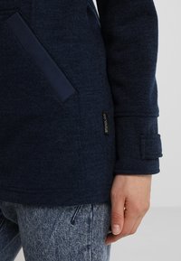 Marinblå fleecejacka med en mjuk textur, utrustad med sidofickor, en logotypetikett och en knappad mudd.