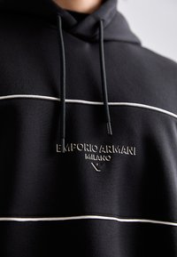 Čierna mikina s kapucňou s bielymi vodorovnými pruhmi a vyšitým logom "EMPORIO ARMANI MILANO" na hrudi, viditeľné čierne šnúrky.