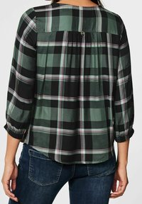 Blouse à carreaux verts et noirs avec manches longues, poignets élastiques et empiècement. Fermeture par boutons au dos, texture de tissu douce.