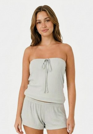 Jeune femme portant un haut bustier côtelé gris clair et un short assorti, debout devant un fond blanc uni, avec un léger sourire.