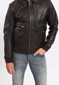 Hnědá kožená bombera s uzavíráním na zip. Má dvě přední kapsy a žebrované manžety. Hladká textura s klasickým límcem.