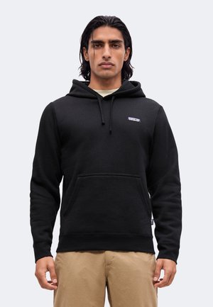 Homme aux cheveux foncés portant un sweat à capuche noir avec poche avant et petit logo, associé à un pantalon beige, debout devant un fond uni.