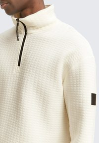 Lichtgekleurde fleece pullover met een verhoogd ruitpatroon, een halve rits kraag en een zwarte accentpatch op de mouw.