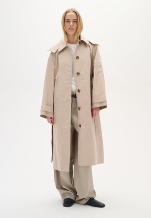 Un trench coat beige con un ampio colletto, chiusura con bottoni visibili e polsini arrotolati. Indossato sopra una camicia bianca e pantaloni beige, con scarpe nere.