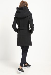 Black coat with a large, folded collar, belted waist, long sleeves, and a tailored fit, paired with dark leggings and athletic shoes.