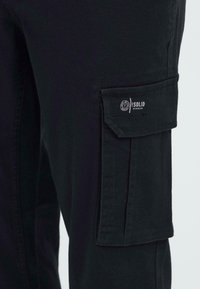 Schwarze Cargo-Hosen aus strapazierfähigem Material verfügen über eine Seitentasche und ein Etikett mit der Aufschrift "ISOLID Denmark". Die Textur ist glatt mit einem matten Finish.