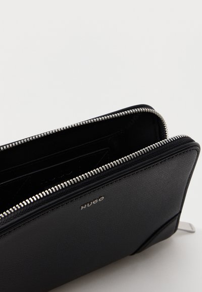 Portefeuille en cuir noir avec fermeture éclair partiellement ouvert, montrant les emplacements pour cartes à l'intérieur, avec une fermeture éclair argentée et le logo "HUGO" embossé à l'avant.