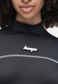 Top preto de gola alta feito de tecido elástico, com costuras em contraste e um patch de logótipo branco no peito.