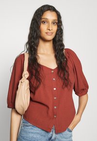 Blusa de cor ferrugem com mangas bufantes, cinco botões à frente e um design em V. Combinada com uma bolsa bege com um detalhe de nó.
