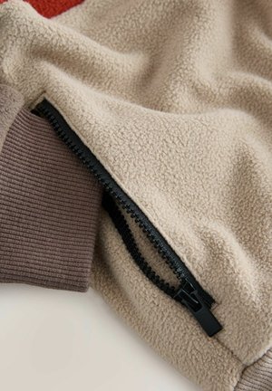 Beżowa tkanina polarowa z czarnym zamkiem błyskawicznym i brązowym ściągaczem na rękawie kurtki lub swetra.