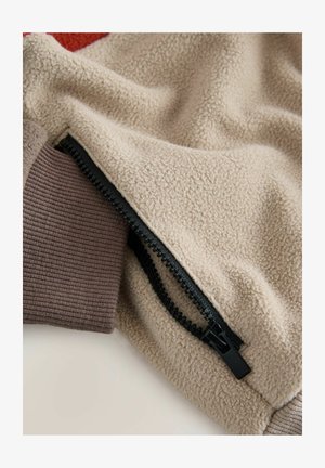 Beige színű bolyhos anyag fekete cipzárral és barna bordás kötött mandzsettával egy dzsekiben vagy pulóver ujján.