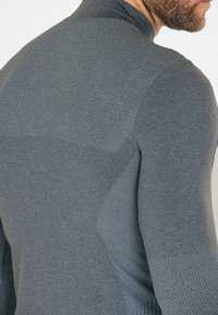 Grå langærmet træningsshirt med tekstureret øverste del, glat nederste del og tætsiddende design. Har diskrete mønsterdetaljer.