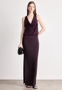 Μακριά, χωρίς μανίκια, έντονα μωβ φόρεμα με ντραπέ λαιμό και λεία υφή, καθώς και πλευρική σχισμή. Το μοντέλο κρατά μια μικρή μαύρη τσάντα.