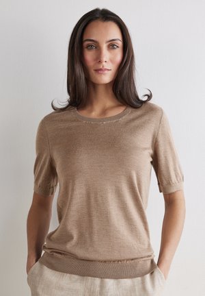 Frau mit langen dunklen Haaren, die einen hellbraunen Kurzarmpullover und beige Hosen trägt, steht vor einer einfarbigen hellen Wand.