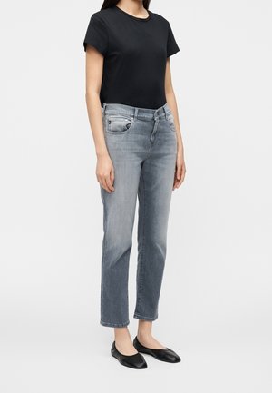 Femme portant un t-shirt noir à manches courtes, un jean droit gris clair et des ballerines noires, debout devant un fond clair uni.
