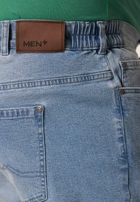 I jeans in denim azzurro chiaro presentano un'etichetta in pelle marrone sulla vita con la scritta "MEN+". Include tasche posteriori con dettagli in bottone argentato.